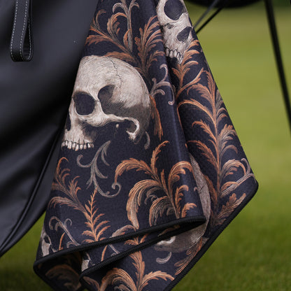 Black golf towel with skull and leaf pattern draped over a black bag on a green background