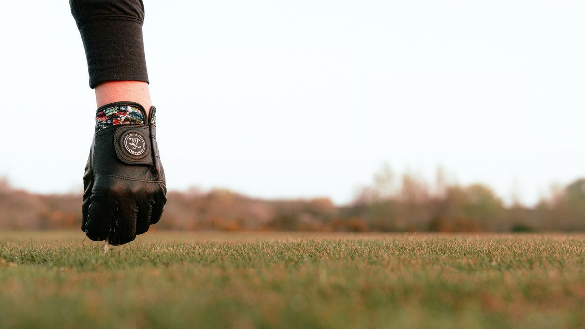 A close-up of a person's hand wearing a black golf glove with a colorful wrist strap, standing on a grassy field under a bright sky.