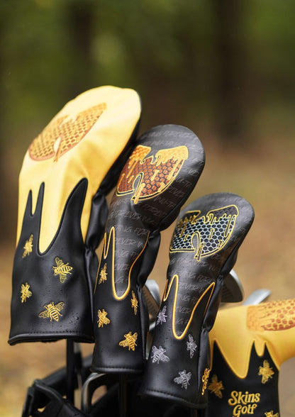 Close-up of golf club headcovers in black and yellow with gold bee and honeycomb embroidery, including a visible 'Skins Golf' logo on one cover, set against a blurred outdoor background.