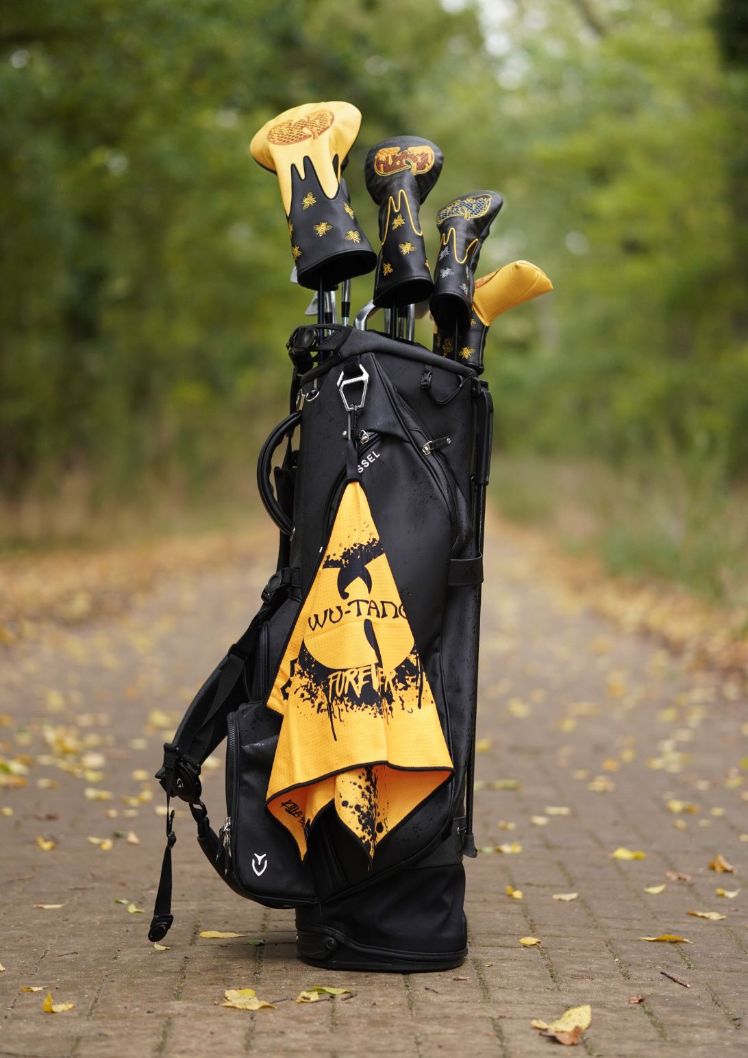 A black golf bag standing on a brick path outdoors, filled with golf clubs that have yellow and black headcovers, with a yellow towel featuring a black graphic hanging from the side.