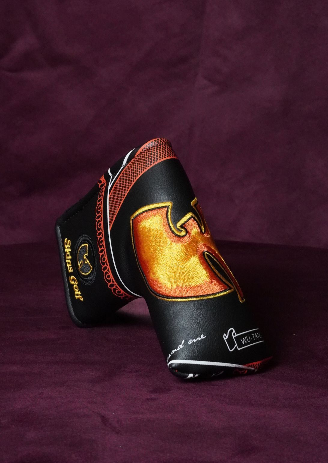 A custom black golf club headcover featuring a large metallic gold and orange flame design with a stylized 'W' logo, resting on a dark purple textured fabric background.