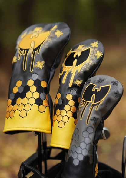 Three black and yellow golf club headcovers with honeycomb and bee graphics, displayed in a golf bag against a blurred outdoor background.