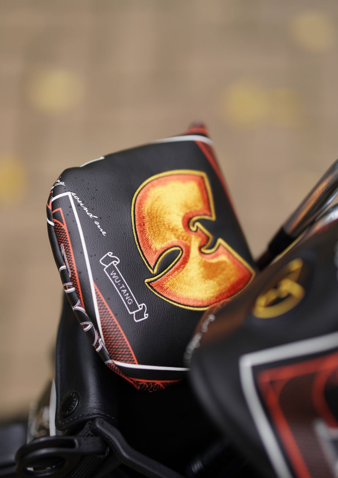 A close-up of a black golf club headcover featuring a large gold and orange Wu-Tang Clan 'W' logo, with red and white graphic details, resting on a golf bag.