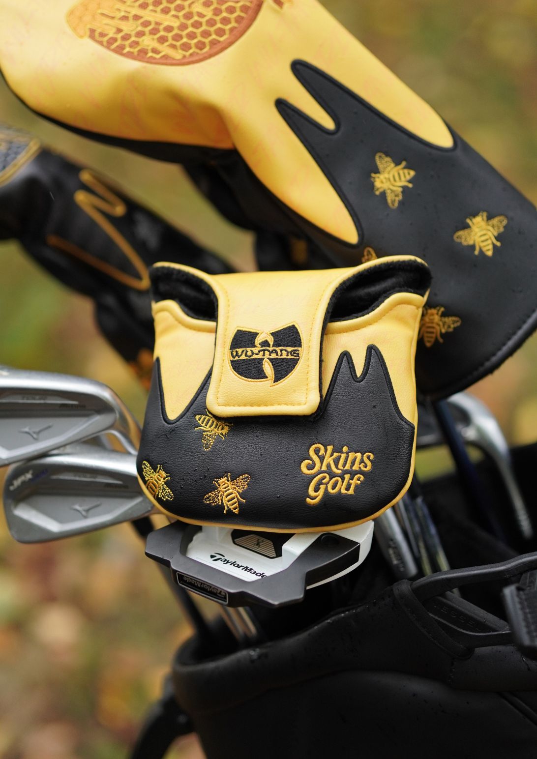 A close-up of a black and yellow golf headcover with gold bee motifs and 'Skins Golf' branding, alongside matching golf headcovers in a bag, set against a blurred outdoor background.