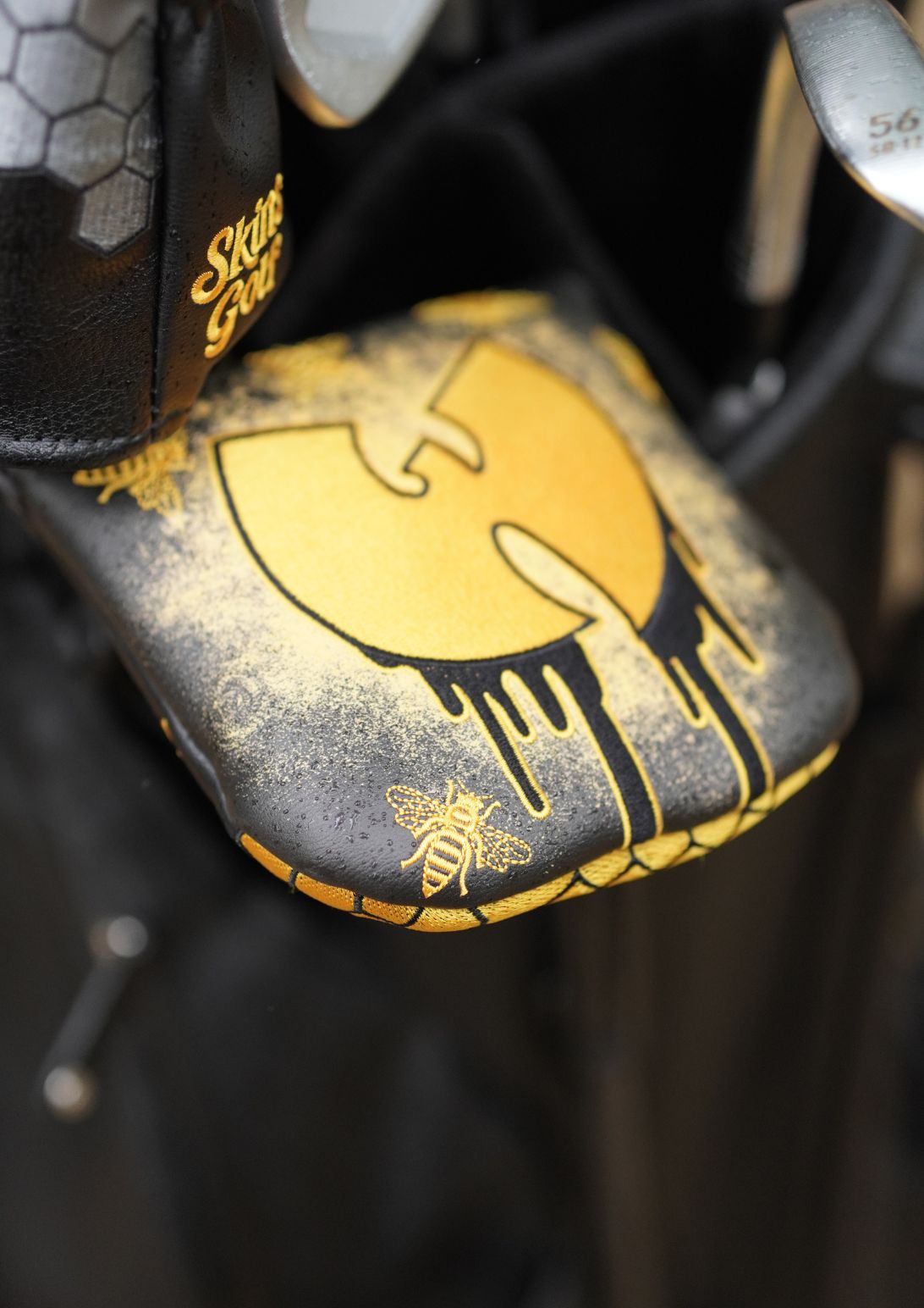 Close-up of a custom black and gold golf club headcover featuring a stylized Wu-Tang Clan-style logo with a bee graphic, shown in a bag.