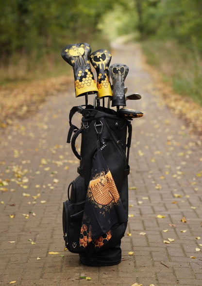 A black golf bag stands on a brick path outdoors, holding several golf clubs with matching black and gold headcovers. The background shows a blurred wooded path with fallen leaves.