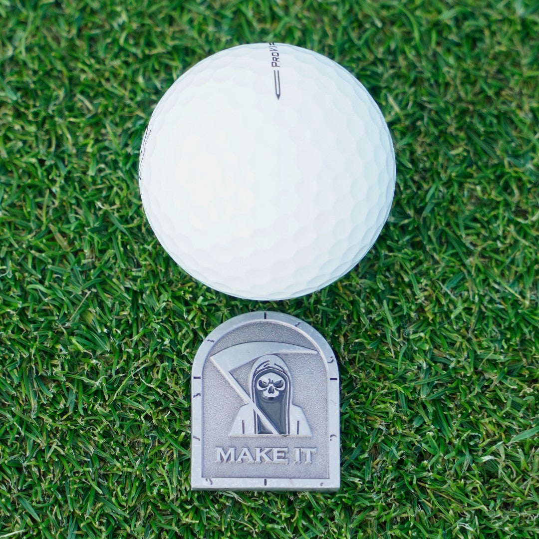 White golf ball on grass with a metal ball marker labelled 'MAKE IT'.