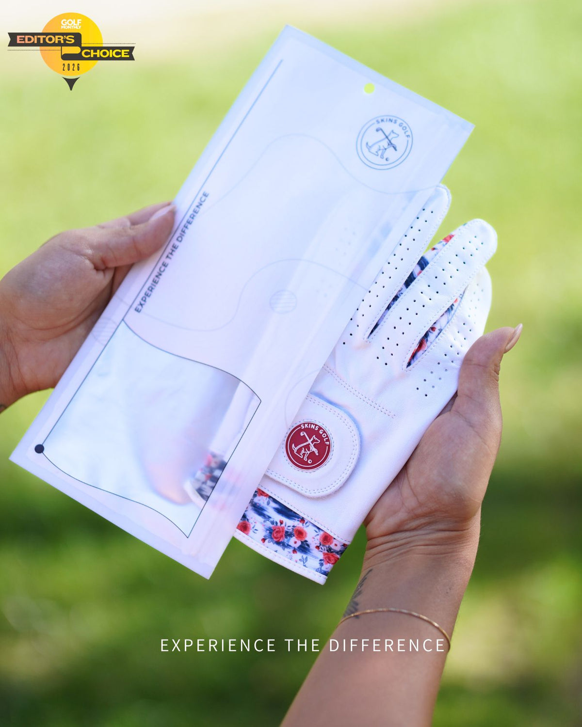 A person holds a white golf glove with a red and white patterned cuff next to its white packaging sleeve, set against a blurred green outdoor background.