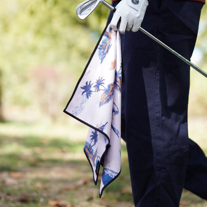 A close-up of a golf club being held by a person wearing dark navy pants and a white glove, with a patterned golf towel featuring blue palm trees and orange accents hanging from the club.
