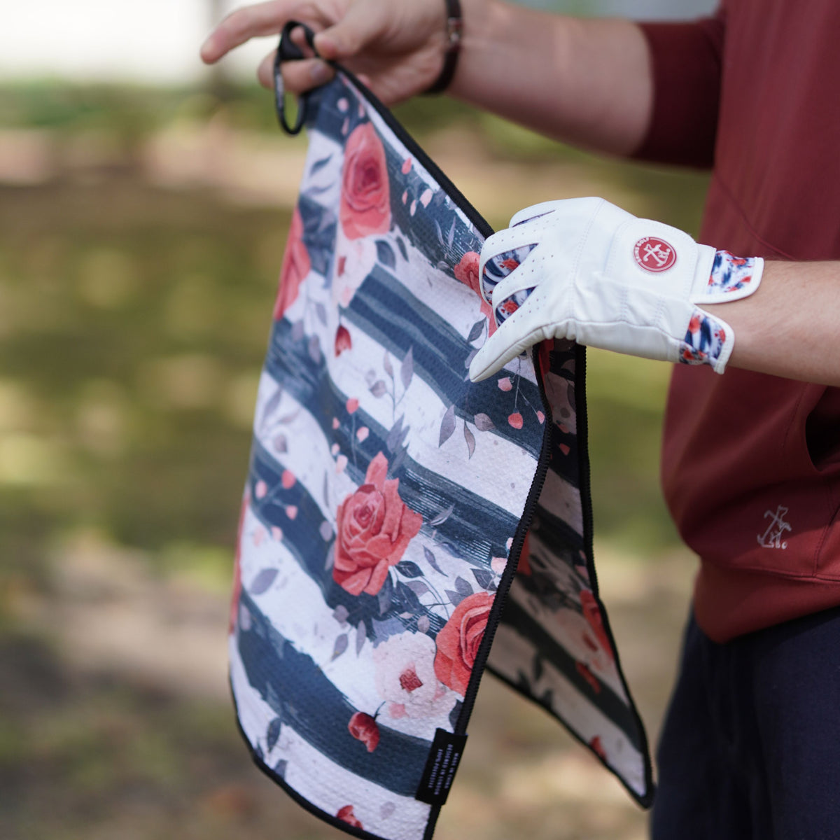 A person outdoors holds a folded golf towel with a navy, white, and red rose pattern. The person is wearing a white golf glove with a red logo.