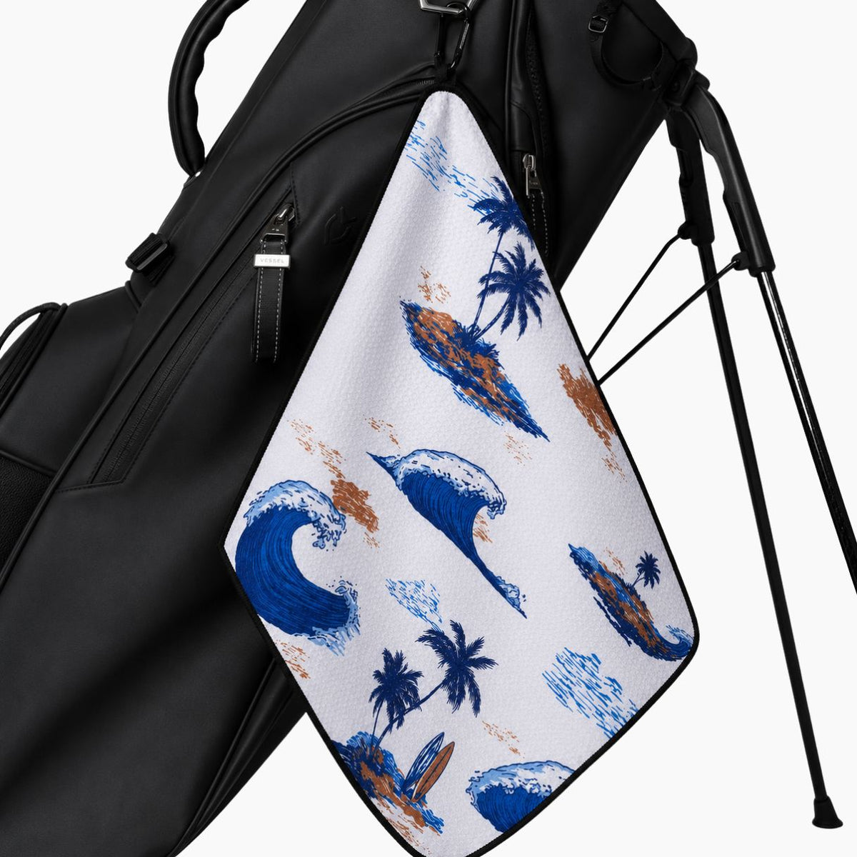 A black golf bag with a white towel hanging from the side pocket. The towel features a blue and brown tropical surf pattern with palm trees and surfboards.